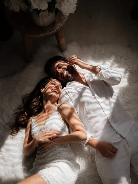 Couple laughing together on a white rug with dramatic side lighting, engagement photos.