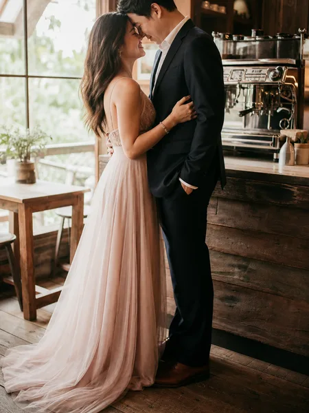 Couple with foreheads touching, standing by a large window in a coffee shop, engagement photos.