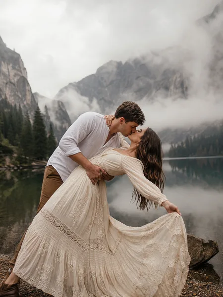 Couple in a gentle dip kiss beside a misty mountain lake, her bohemian dress flowing, with reflection, engagement photos.