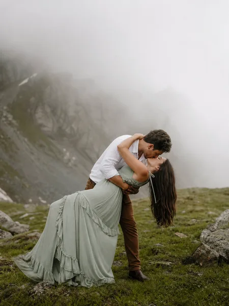 Couple kissing in a dip pose in misty alpine meadow, bohemian dress, engagement photos.