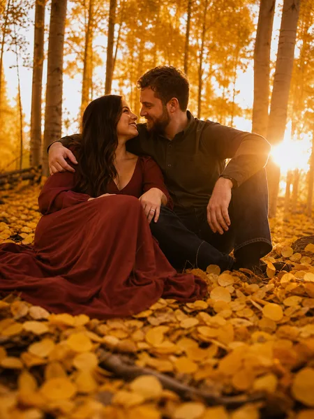Couple sitting closely on fallen aspen leaves in golden hour light, burgundy dress, engagement photos