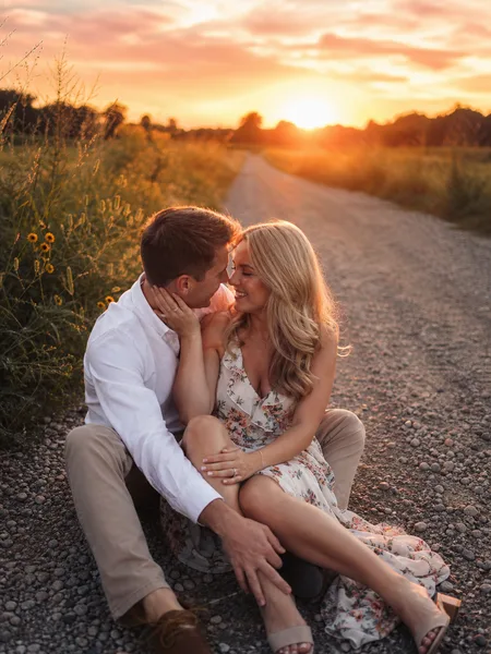 Couple sitting together on a country road at sunset, floral dress, wildflowers, warm light, romantic engagement photos.