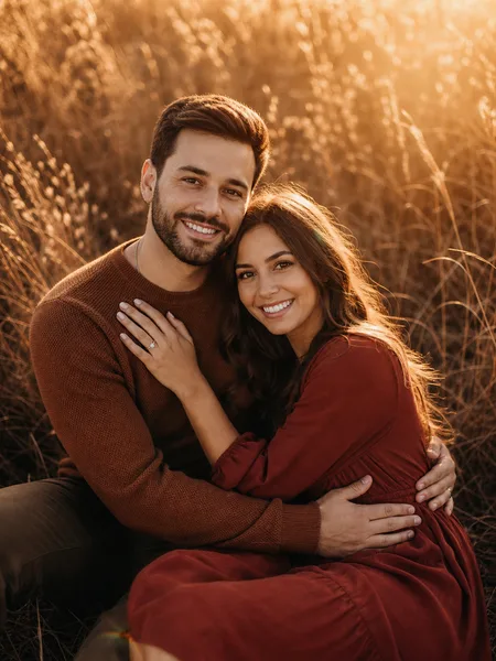 Couple sitting closely in a dried grass field during golden hour, romantic engagement photos.