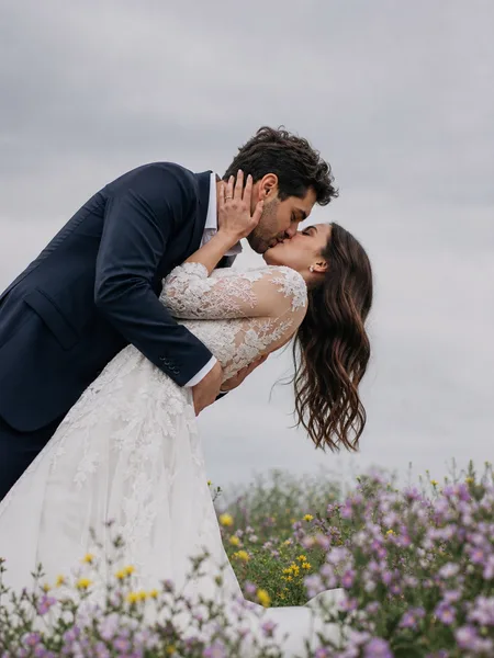Couple shares a gentle dip kiss in a wildflower meadow on an overcast day, engagement photos with a white dress.