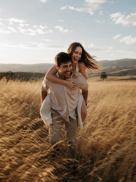 Couple enjoying a piggyback ride in a vast dried grass field, editorial engagement photos.