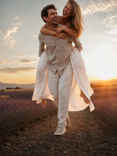 Couple piggyback ride in lavender field at golden hour, white dress flowing, joyful romantic moment engagement photos