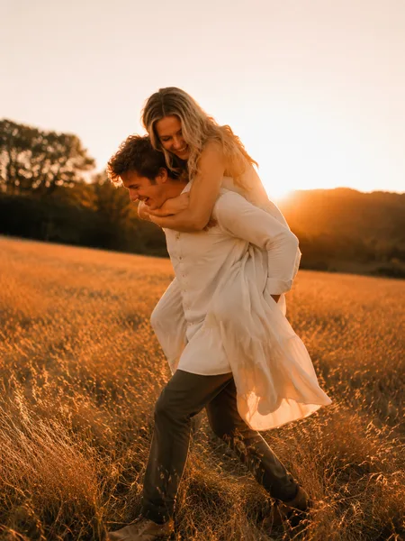 Couple piggyback ride in tall grass prairie at golden hour, engagement photos.