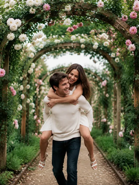 Groom giving bride piggyback ride under rose-covered arches in an editorial style, soft light, engagement photos.