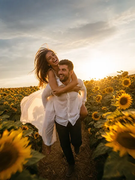 A couple piggybacking through a sunflower field in golden hour, her white linen dress flowing, backlit sun, engagement photos.