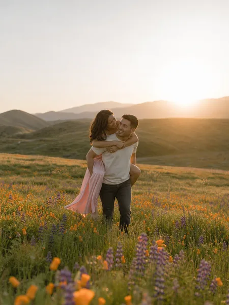 Couple gives a piggyback ride in a wildflower meadow at sunrise, engagement photos capturing laughter and soft golden light.