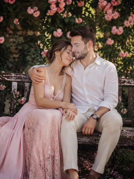 Couple sitting closely on a bench in a blooming rose garden, soft morning light, for their engagement photos.