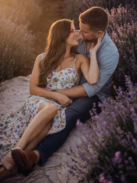 Couple sitting close on a blanket in lavender field at sunset, floral dress, warm golden light engagement photos