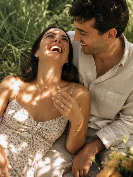 Couple laughing together on picnic blanket in park, she in floral dress, golden light, engagement photos.