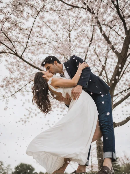 Couple in a dip kiss under cherry blossoms, overcast light, white dress, engagement photos.
