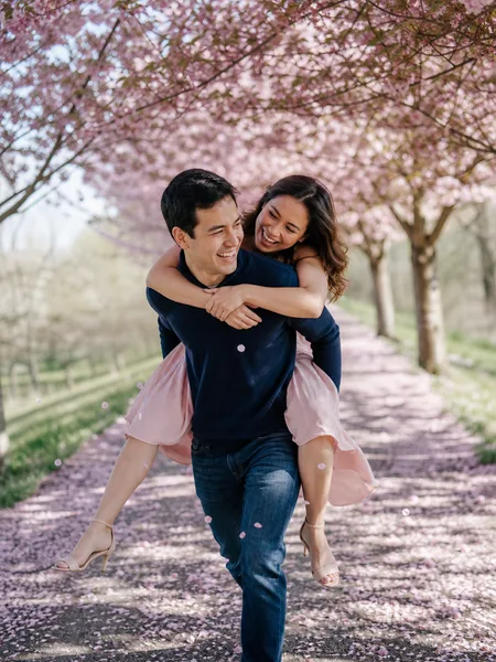 Couple piggyback riding on a cherry blossom path, morning light, pink dress, engagement photos.