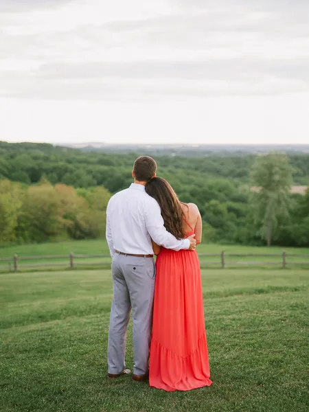 Couple from behind watching horizon in park, bright dress, overcast sky, engagement photos