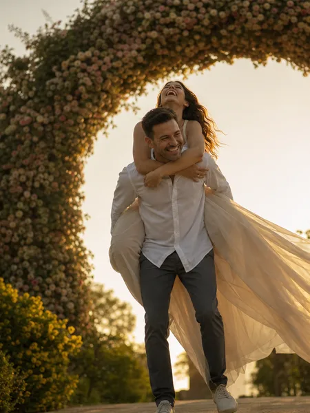Piggyback ride under a flower arch in golden light, white flowy dress, engagement photos.