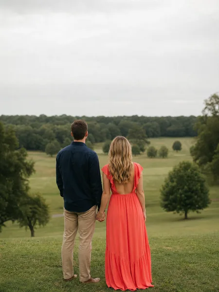 Couple standing in park gazing at horizon, she in bright coral dress, overcast sky, engagement photos.