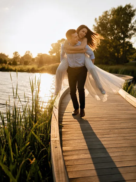 Piggyback ride on wooden bridge at golden hour, white dress flowing, couple laughing, pond with cattails, engagement photos.