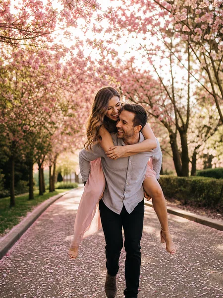 Groom-to-be giving his partner a piggyback ride along a cherry blossom path, both smiling in morning light, engagement photos.