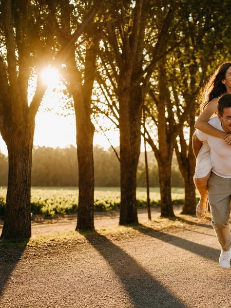 A couple piggyback riding on a sunlit tree-lined avenue at golden hour, engagement photos.
