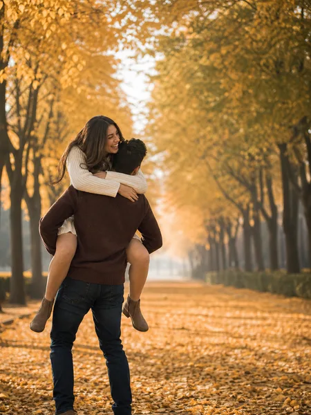 Man giving woman a piggyback ride on a tree-lined park avenue covered in golden fall leaves, soft light, engagement photos.