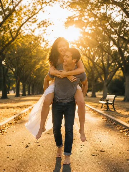 Man carrying woman piggyback on a park path at sunset, her white dress flowing, engagement photos.
