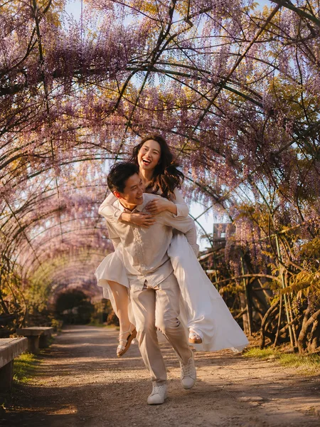 Piggyback ride under blooming wisteria arch, white flowy dress, golden hour light, engagement photos.