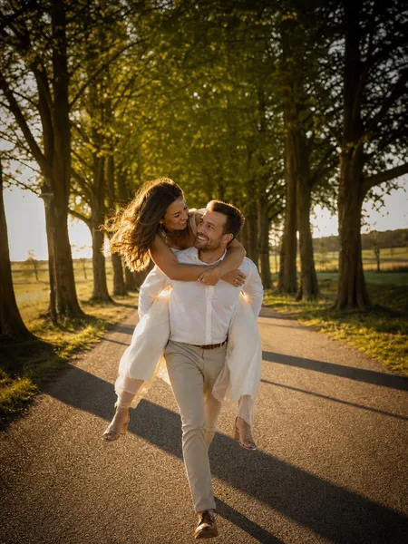 Couple piggyback riding on a tree-lined avenue at golden hour, engagement photos