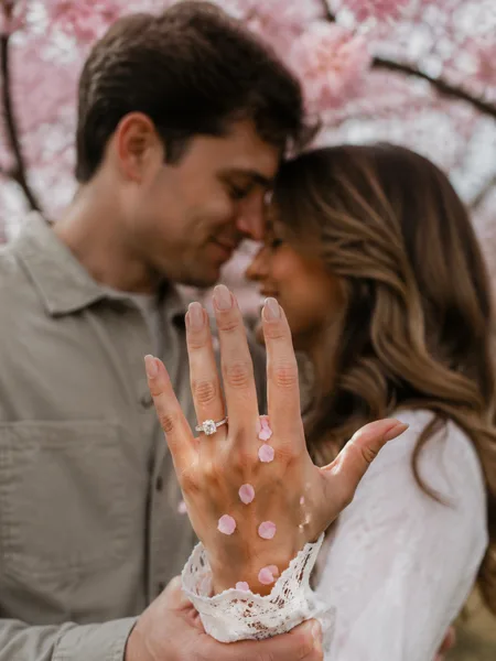 Close-up of an engagement ring on a hand with cherry blossom petals, white dress, overcast light, engagement photos.