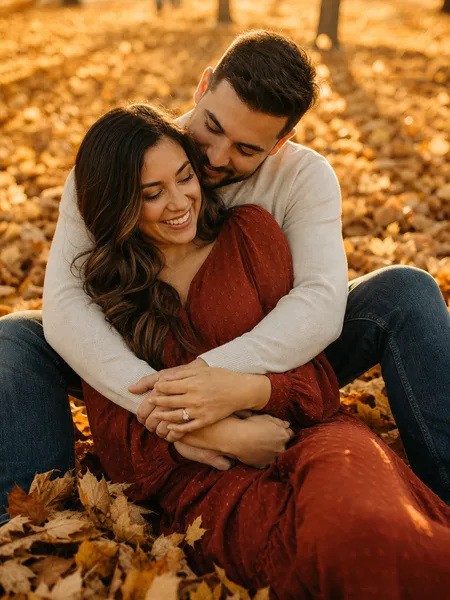 Couple sitting closely on a bed of golden leaves under a tree-lined avenue, warm sunset light, engagement photos.